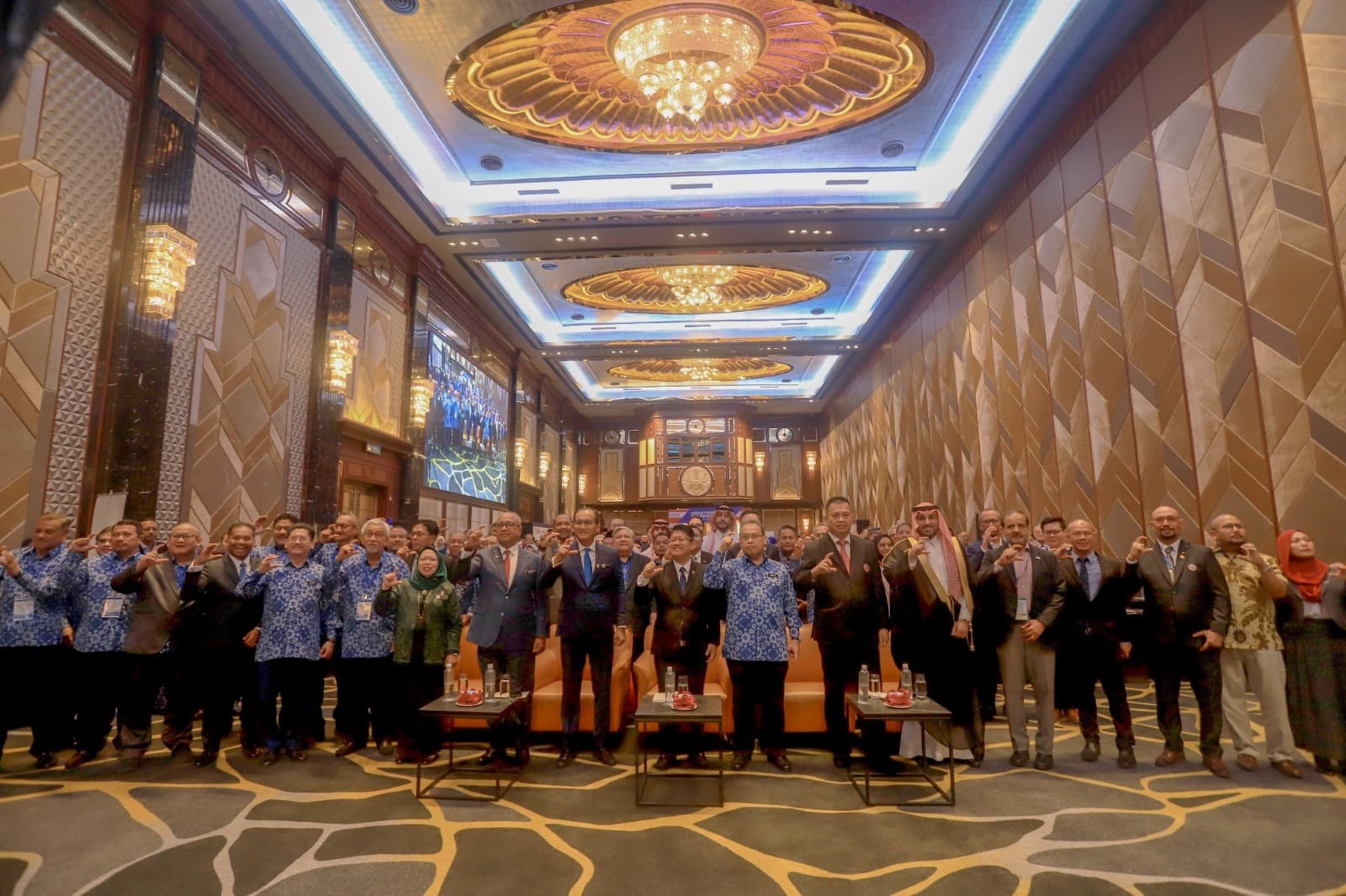 A large group of delegates in suits and batik standing together for a photograph in a hotel ballroom.