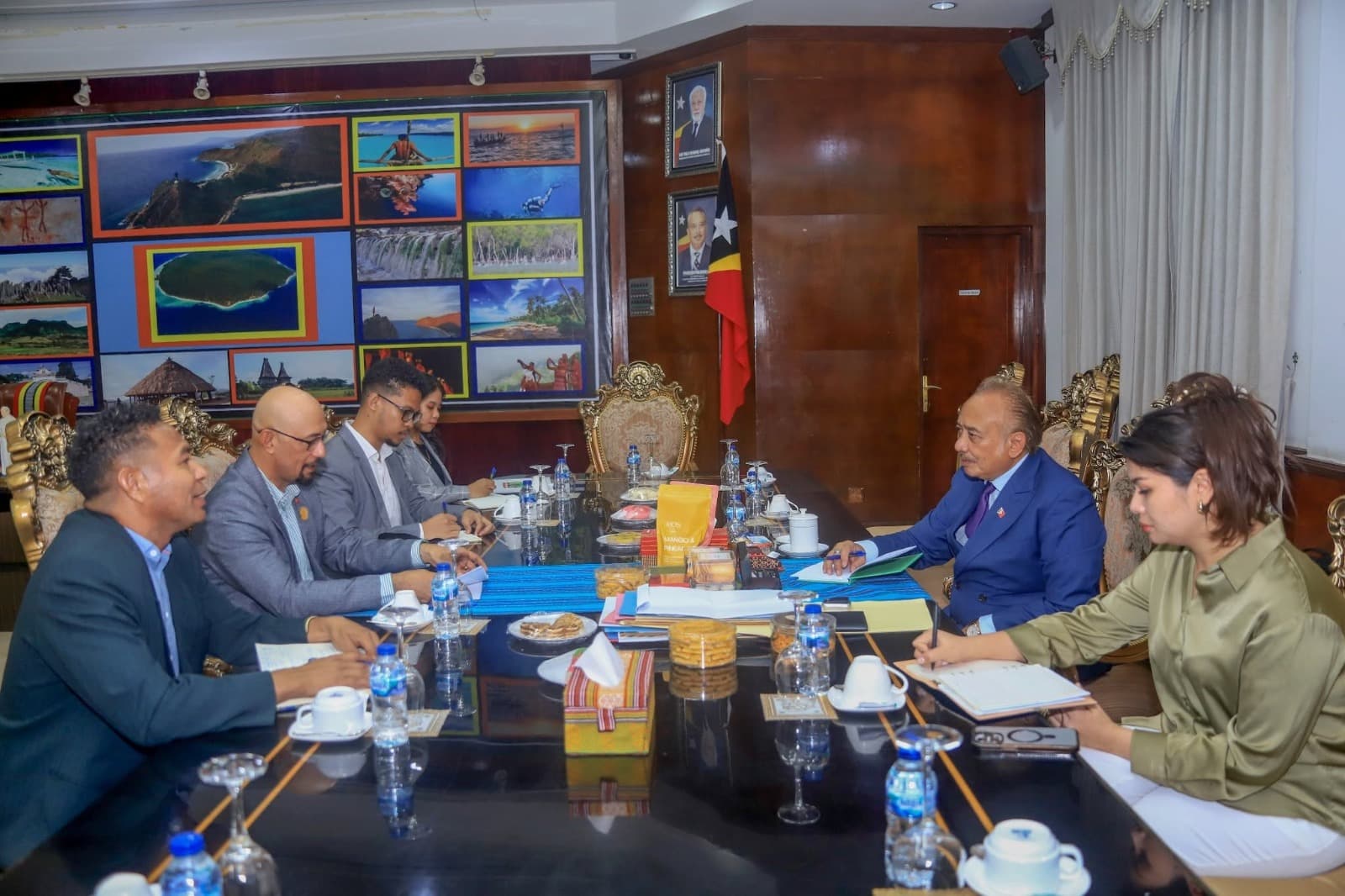 Officials seated on both sides of a meeting table in a government office, with the flag of Timor-Leste and official portraits on the wall behind.
