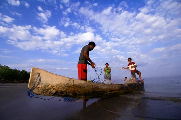 Three people hauling a net from a wooden outrigger canoe drawn up on a beach under a wide cloudy sky.
