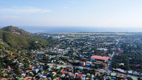Aerial view of a coastal town of brightly roofed houses between a green hill and the sea.