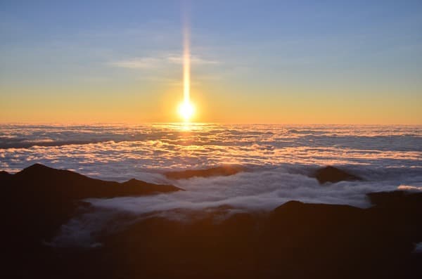 The sun rising over an unbroken sea of cloud, seen from a high ridge with dark peaks in front.