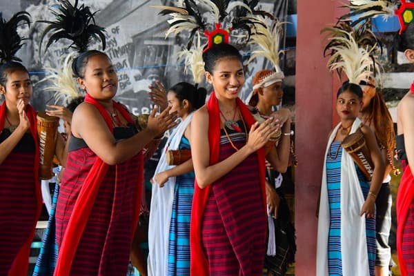 Young women in red and striped tais cloth with feather headdresses, clapping during a performance.