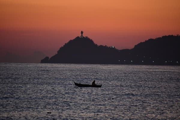 A lone fisherman in a small boat on calm water at sunset, a headland silhouetted behind.